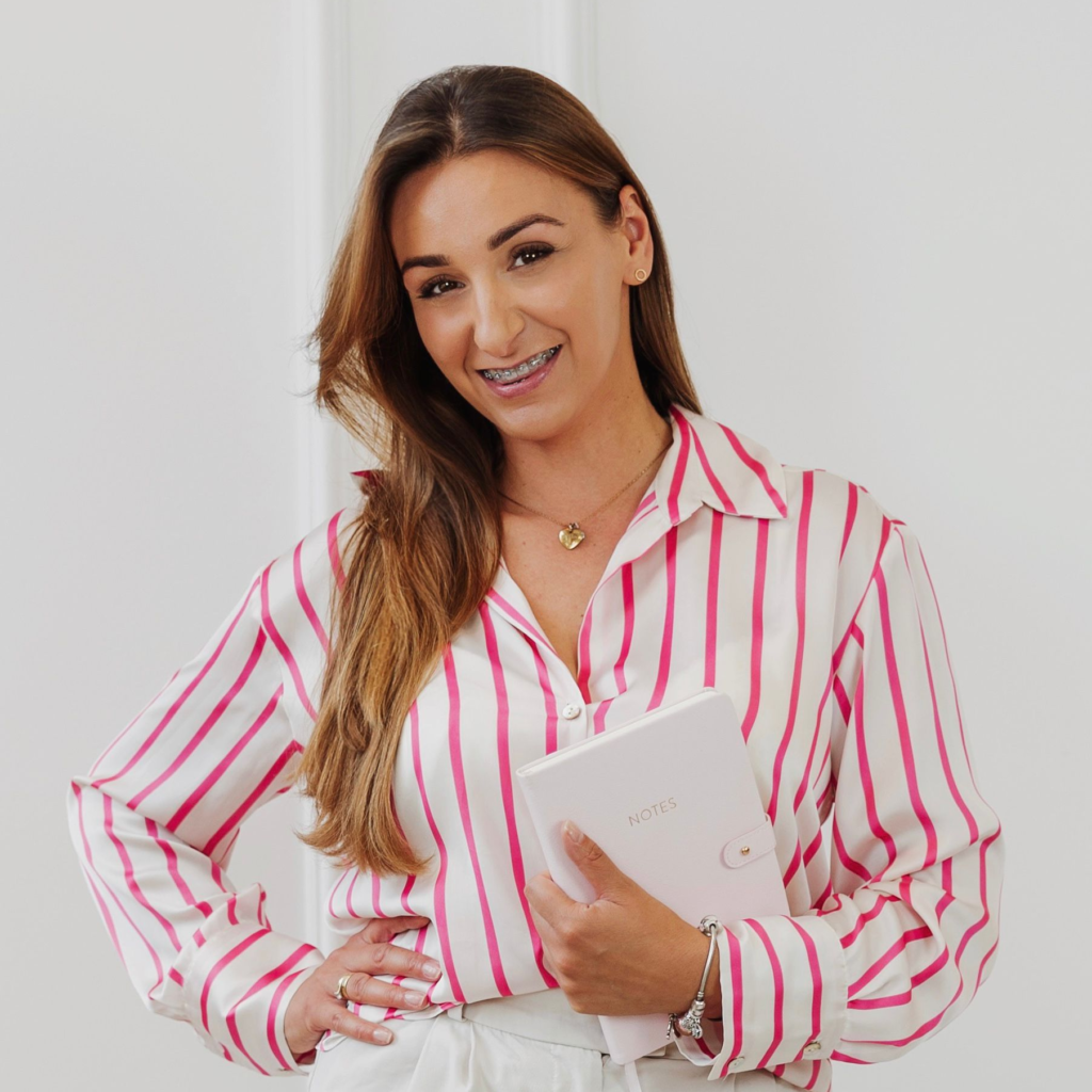 Uśmiechnięta młoda kobieta w biało-różowej koszuli w paski, trzymająca różowy notes z napisem "NOTES".