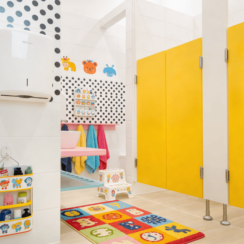 Jasna łazienka przedszkolna z żółtymi drzwiami kabin, kolorowym dywanikiem z motywami zwierząt oraz akcesoriami dostosowanymi do wzrostu dzieci.