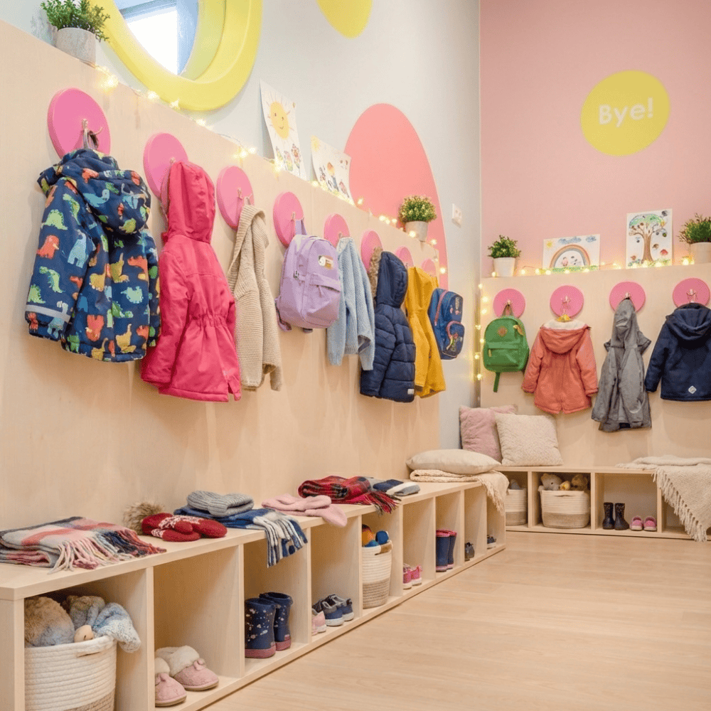 Uporządkowana szatnia przedszkolna z jasnymi drewnianymi szafkami na buty, kolorowymi kurtkami na różowych wieszakach i plecakami dziecięcymi